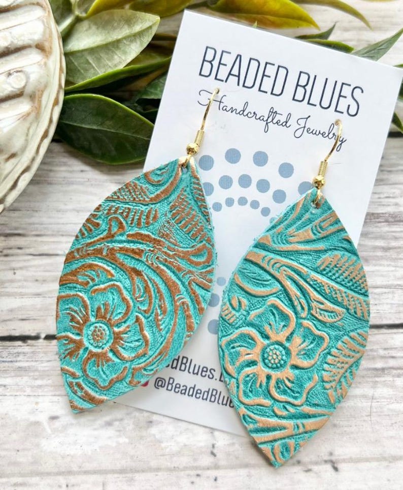 May include: Teal leather earrings with a gold embossed floral pattern. The earrings are shaped like a teardrop and have a gold hook closure. The earrings are handmade and are a beautiful statement piece.