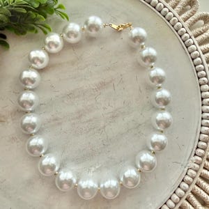 May include: A necklace composed of large, round, white faux pearls strung together with small gold-toned metal spacers between each bead. The necklace features a gold-toned metal lobster claw clasp closure at the top, completing the circular design.