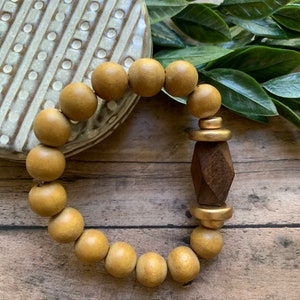 May include: A wooden beaded bracelet with a gold accent and a geometric wooden charm. The bracelet is on a wooden surface.