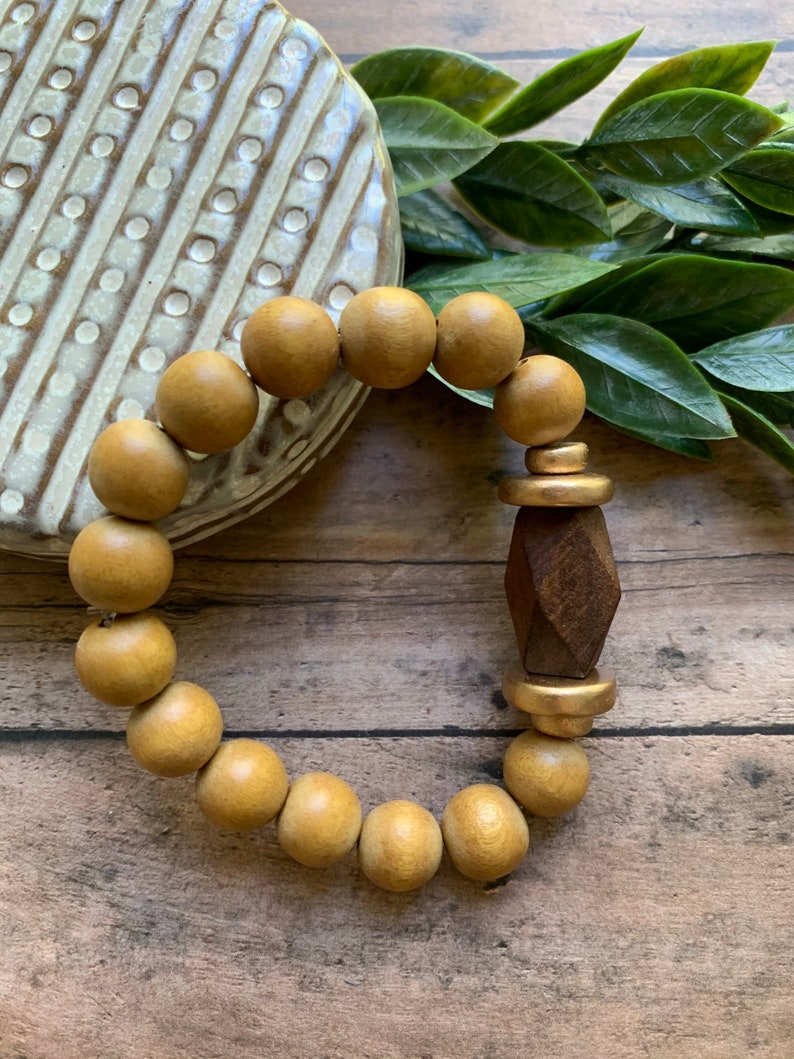 May include: A wooden beaded bracelet with a gold accent and a geometric wooden charm. The bracelet is on a wooden surface.