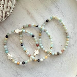 May include: Three beaded bracelets with a mix of pastel-colored beads, including light blue, green, and peach. Each bracelet features a white cross charm and gold-colored accents. The bracelets are displayed on a white wooden tray.
