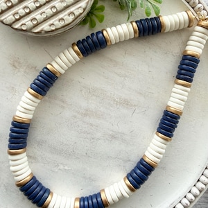 May include: A chunky necklace featuring alternating white, navy blue, and gold-colored disc beads. The necklace is displayed on a white, textured surface, showcasing its layered design and nautical color scheme. The necklace is a statement piece.