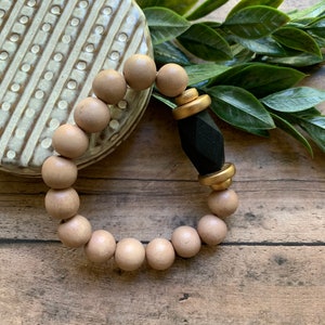 May include: A wooden bead bracelet with a black and gold accent. The bracelet is made of natural wood beads and has a geometric black charm with gold accents.