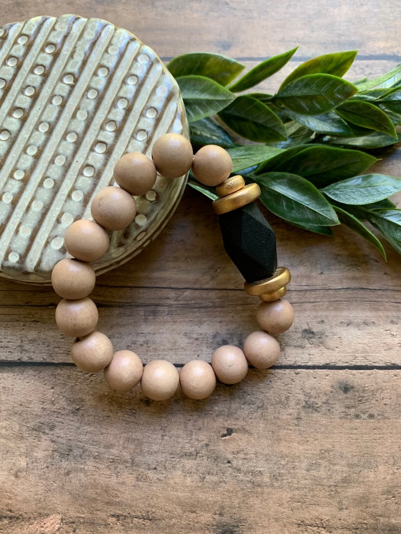May include: A wooden bead bracelet with a black and gold accent. The bracelet is made of natural wood beads and has a geometric black charm with gold accents.