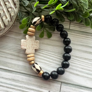 May include: A black and white beaded bracelet with a white cross charm. The bracelet is made with wooden beads and has gold accents.