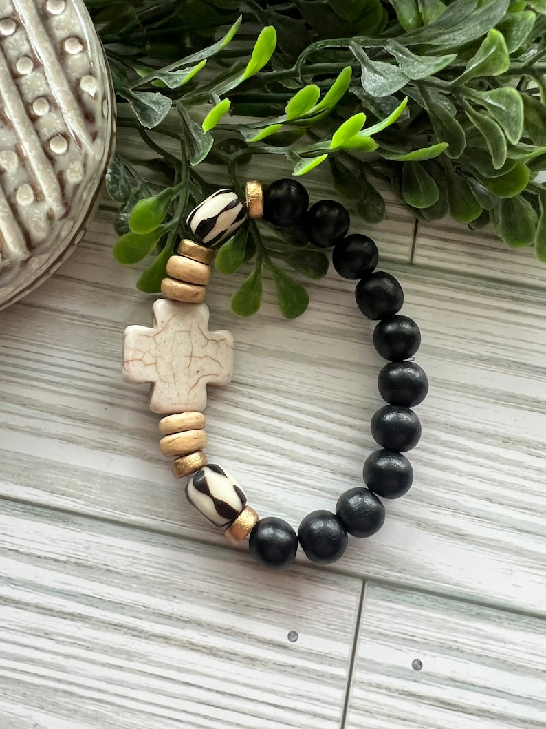 May include: A black and white beaded bracelet with a white cross charm. The bracelet is made with wooden beads and has gold accents.
