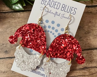 May include: Red and white glitter Santa Claus earrings with gold hooks. The earrings are handmade and feature a red glitter Santa hat and a white glitter beard. The earrings are labeled "Beaded Blues Handcrafted Jewelry".