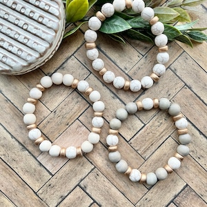 May include: Three beaded bracelets with white, gray, and cream-colored beads. The bracelets have gold-colored spacers and are arranged on a wooden surface with a decorative ceramic piece and greenery in the background.