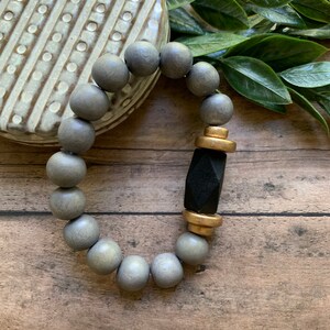 May include: A gray wooden beaded bracelet with a black and gold accent. The bracelet is on a wooden surface.