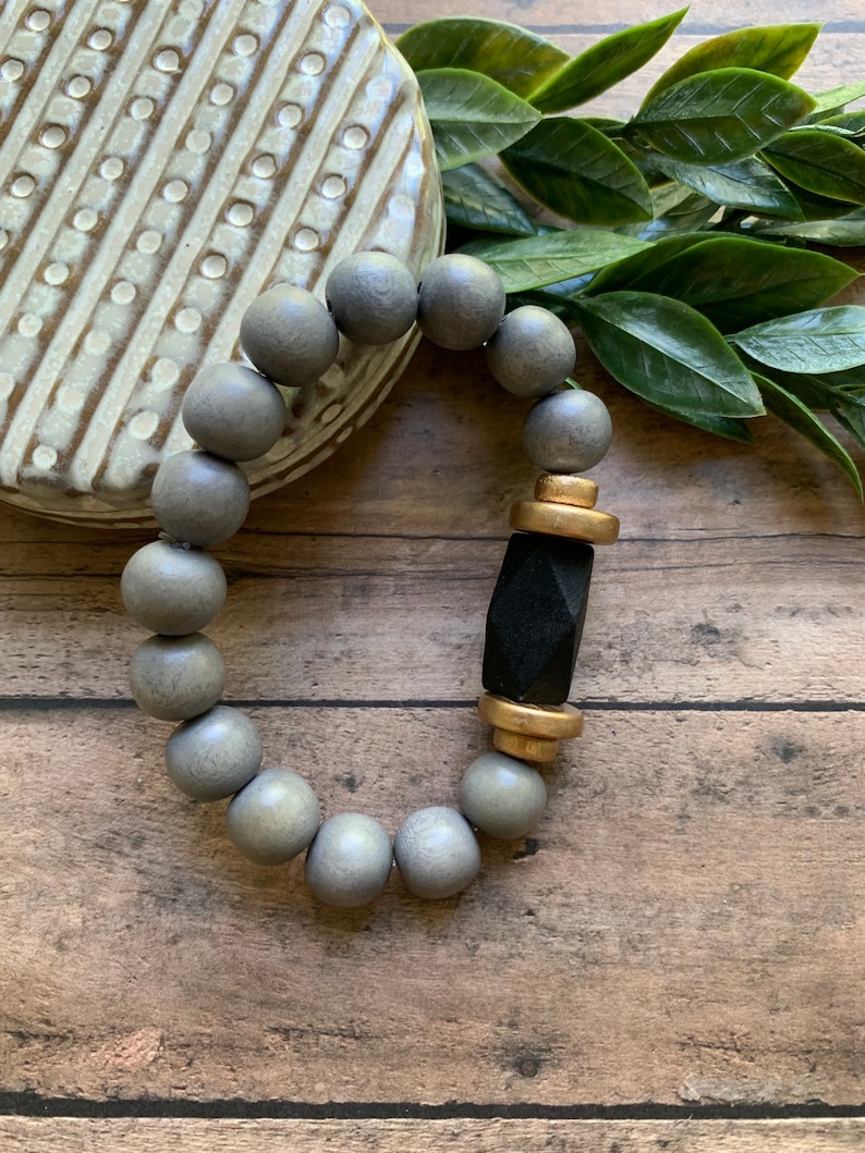 May include: A gray wooden beaded bracelet with a black and gold accent. The bracelet is on a wooden surface.