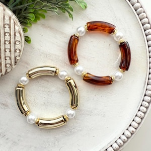 May include: Two beaded bracelets. One bracelet features gold-colored, cylindrical beads and white faux pearls. The other bracelet has amber-colored, cylindrical beads and white faux pearls. Both bracelets are on a white, decorative tray.