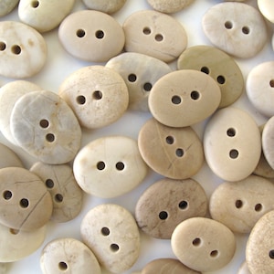 May include: A collection of oval-shaped, natural stone buttons in various shades of beige and brown. The buttons have two holes each and are arranged in a random pattern on a white background.