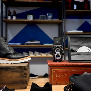 Puede incluir: Bota de cuero negro con suela blanca y ribete marr&oacute;n, expuesta sobre una caja de madera. Una c&aacute;mara vintage y una fotograf&iacute;a en blanco y negro enmarcada est&aacute;n sobre un cofre rojo. Estanter&iacute;as en el fondo contienen varios art&iacute;culos.