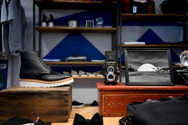 Puede incluir: Bota de cuero negro con suela blanca y ribete marr&oacute;n, expuesta sobre una caja de madera. Una c&aacute;mara vintage y una fotograf&iacute;a en blanco y negro enmarcada est&aacute;n sobre un cofre rojo. Estanter&iacute;as en el fondo contienen varios art&iacute;culos.