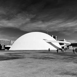 Puede incluir: Fotograf&iacute;a en blanco y negro de un gran edificio blanco en forma de c&uacute;pula con una pasarela curva. El edificio se alza sobre un cielo nublado, con gente caminando en primer plano.