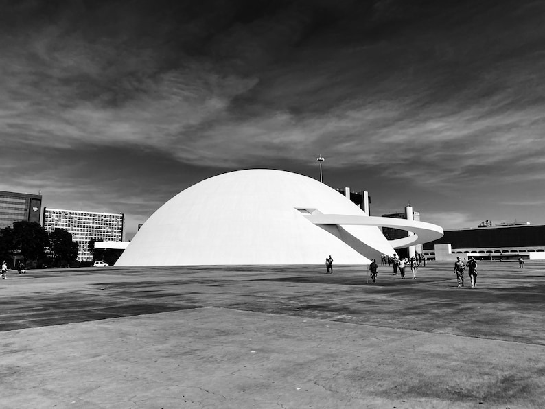 Puede incluir: Fotograf&iacute;a en blanco y negro de un gran edificio blanco en forma de c&uacute;pula con una pasarela curva. El edificio se alza sobre un cielo nublado, con gente caminando en primer plano.