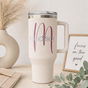 May include: Cream-colored insulated tumbler with a handle, a silver lid, and a clear straw. The tumbler features a large magenta letter "M" with the name "Madison" in silver script. A framed print with the text "focus on the good" is in the background.