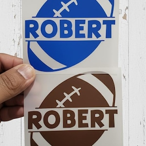 May include: Two football-shaped decals, one blue and one brown, each with the name "ROBERT" in bold letters. The blue decal has white stripes, while the brown one has tan stripes. The decals are held up against a white wooden background.