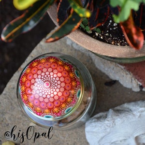 May include: A glass jar with a colourful mandala painted on the lid. The lid is red, orange, yellow, pink, and white with a blue and green border. The jar is sitting on a grey surface. @hisOpal