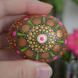 May include: A hand-painted rock with a mandala design in shades of brown, gold, white, pink, and green.
