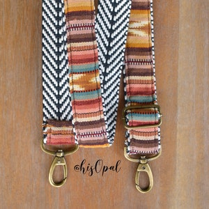 May include: Two patterned bag straps with brass-coloured hardware. One strap features a black and white chevron design, while the other has a colourful woven pattern in shades of orange, brown, and red. The straps are displayed on a wooden surface.