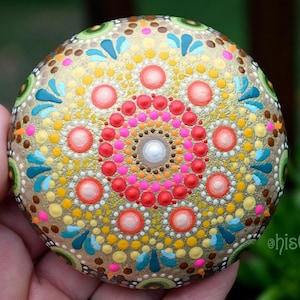 May include: A hand-painted rock featuring a colourful mandala design. The mandala has a central circle with concentric rings of red, pink, orange, yellow and white dots. The outer edge of the rock is decorated with green, brown and blue accents. The rock is painted on a gold background.