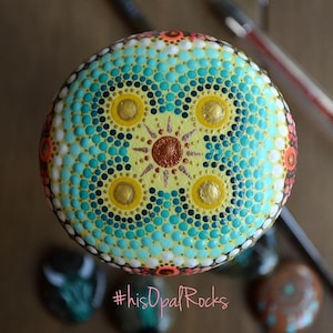 May include: A round, painted rock with a mandala design in shades of yellow, blue, green, and gold. The rock is surrounded by other painted rocks. The hashtag #hisOpalRocks is visible in the bottom of the image.