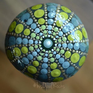 May include: A round, painted rock with a mandala design in shades of green, blue, and silver. The rock is painted with dots and circles, creating a geometric pattern. #hisOpalRocks