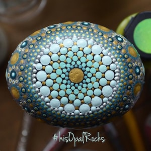 May include: A round, grey stone painted with a mandala design in shades of teal, white, and gold. The stone is surrounded by paint bottles.