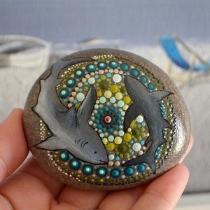 May include: A hand-painted rock with a mandala design featuring two grey sharks and a colourful dot pattern in shades of green, blue, and white.