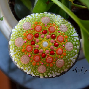 May include: A green, orange, pink and red painted rock with a mandala design. The rock is round and has a glossy finish.