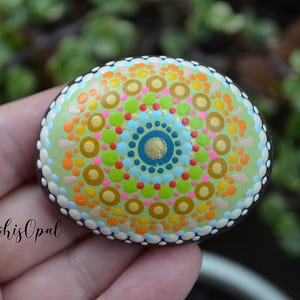 May include: A hand-painted rock featuring a colourful mandala design. The mandala incorporates a range of colours, including green, blue, yellow, orange, pink, and white. The rock is painted with a black outline and has a glossy finish.