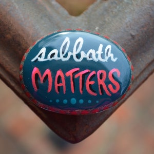 May include: A teal blue painted rock with the words "sabbath matters" in white and pink lettering. The rock has a red border and a few teal blue dots.