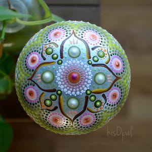 May include: A round, green, white, pink, and brown painted rock with a mandala design. The rock is decorated with dots and swirls.