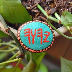May include: A teal painted rock with a white border and red painted symbols. The rock is surrounded by green leaves.