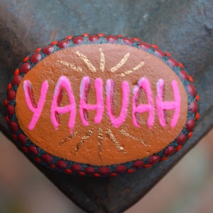 May include: A painted rock with a red and black border and gold accents. The word "YAHUAH" is written in pink letters on the rock.