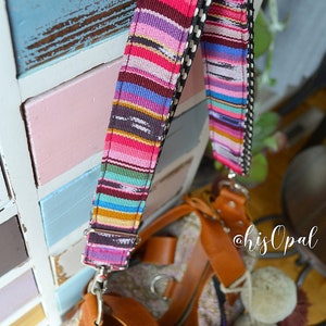 May include: A colourful striped fabric bag with a brown leather strap and a zip closure. The bag has a unique woven design and is accented with fluffy pom-poms. The strap is attached to the bag with a silver metal hook. The bag is hanging on a wooden surface with a painted blue and pink design. The text "@hisOpal" is visible in the image.