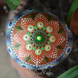May include: A round, painted rock with a mandala design. The mandala features a green, pink, and white centre surrounded by brown leaves and blue and white circles. The rock is painted with a variety of colours, including brown, pink, green, blue, white, and gold. The rock is sitting on a green leaf. #thisopalrocks