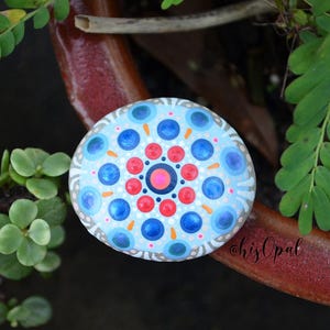 May include: A painted rock featuring a blue, red and white mandala design. The rock is resting on a brown surface with green plants.