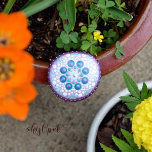 May include: A purple, white and blue mandala painted on a round rock. The rock is sitting on top of a terracotta flower pot.