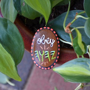 May include: A brown painted rock with white and green lettering that says "obey yayz" with orange and pink polka dots around the edge.