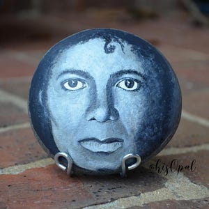May include: A hand-painted, round stone with a portrait in grey and white. The artwork depicts a face with detailed eyes, nose, and lips. The stone rests on a silver metal stand. The artist's signature is visible.
