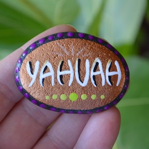 May include: A painted rock with a copper-coloured background and a black outline. The rock has a purple dotted border and the word "YAHUAH" written in white letters with black outlines.