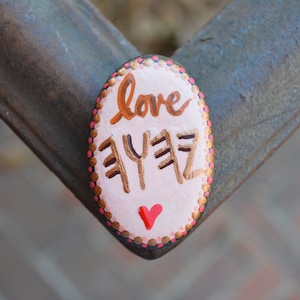 May include: A pink painted rock with gold lettering that says "love aye aye". The rock is decorated with small pink and gold dots around the edge and has a small pink heart painted on the bottom.