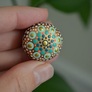 May include: A small, round, white stone painted with a mandala design. The mandala features concentric circles of teal, gold, white, and coral dots.