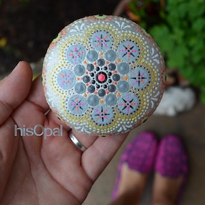May include: A hand is holding a grey stone painted with a mandala design. The mandala features a central circle with a pink dot, surrounded by concentric circles of grey, white and yellow dots. The outer edge of the mandala is decorated with a repeating pattern of white and yellow dots.