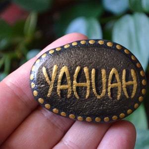 May include: A black painted rock with a gold border and the word "YAHUAH" written in gold paint.