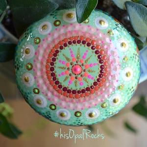 May include: A round, painted rock with a mandala design in shades of pink, green, red, and gold. The rock is surrounded by green leaves. #hisOpalRocks