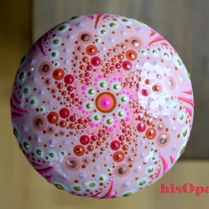 May include: A pink, white and gold painted rock with a mandala design. The mandala features a central circle with a pink dot and gold dots surrounding it. The design is surrounded by concentric circles of pink, white and gold dots.