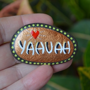 May include: A painted rock with a copper-coloured background and a green and black dotted border. The rock has the word "YAHUAH" written in white with black outlines and a red heart above the word.